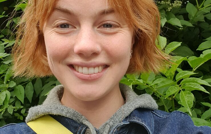 Mary, Nature and Wellbeing Officer smiling at the camera stood in front of trees wearing a denim jacket and yellow bag