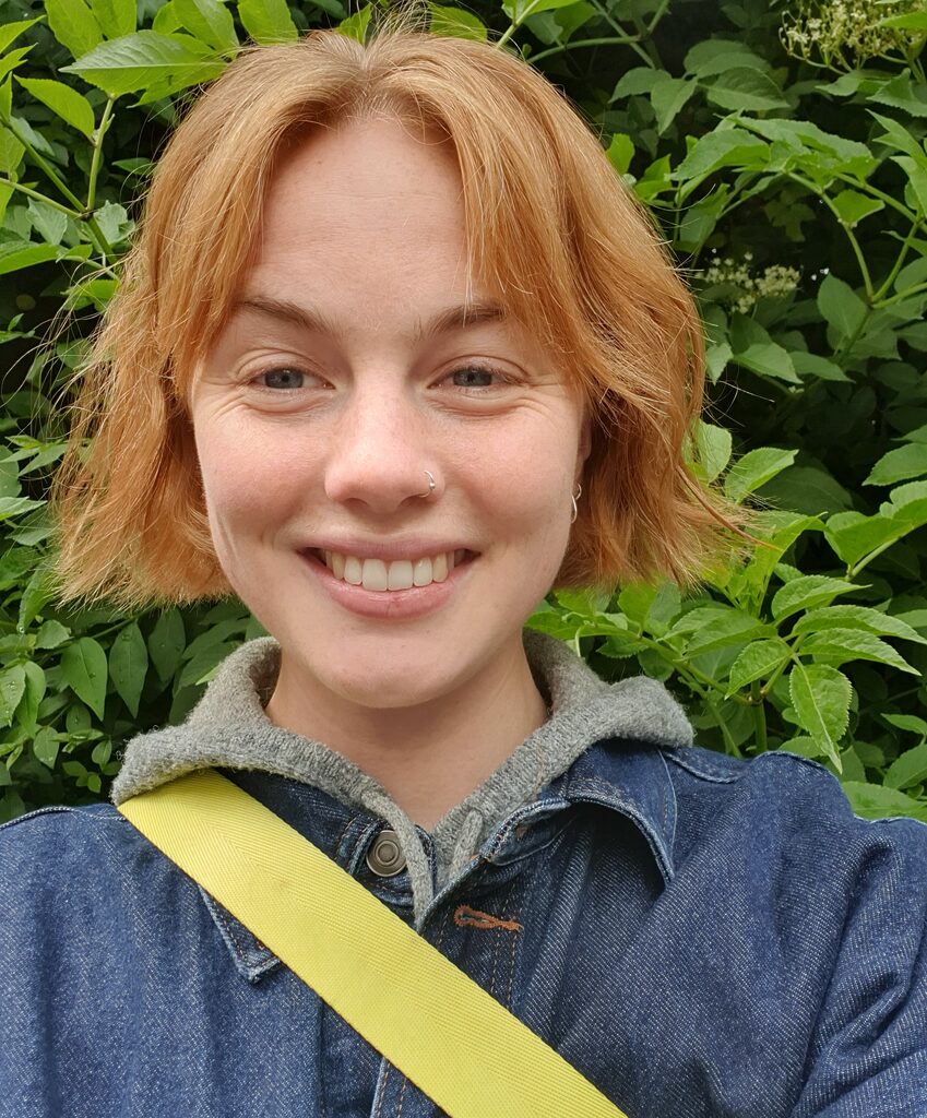 Mary, Nature and Wellbeing Officer smiling at the camera stood in front of trees wearing a denim jacket and yellow bag